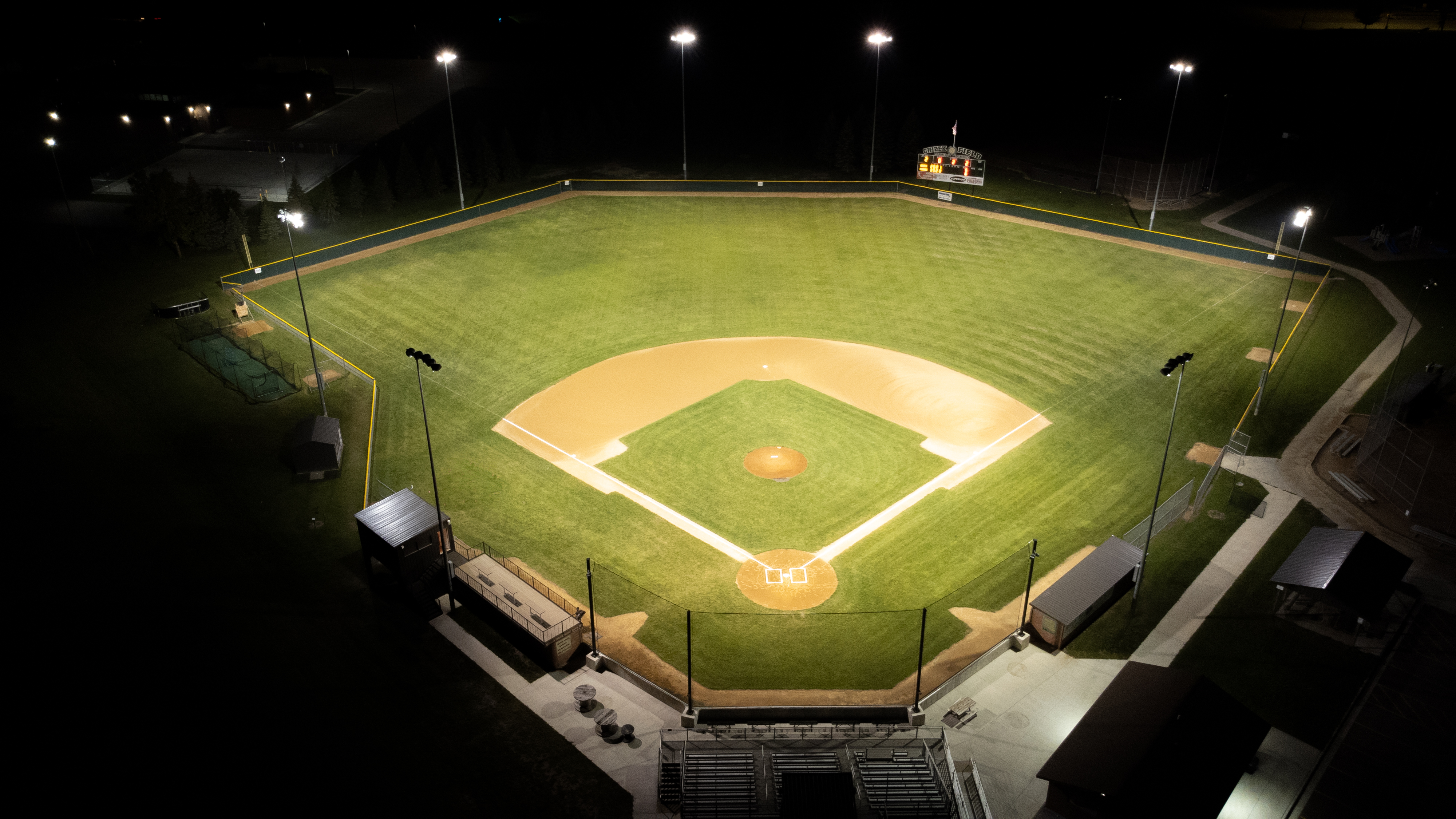 Aerial nighttime view of a brightly lit baseball stadium with a glowing infield, green outfield, bleachers, and surrounding walkways visible against the dark landscape.